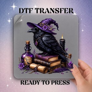 May include: A DTF transfer sheet featuring a black raven wearing a purple witch hat with flowers, perched on a stack of books. The design includes a purple bottle labeled Raven's Brew, a lit black candle, a scroll, and a small vial of purple crystals.