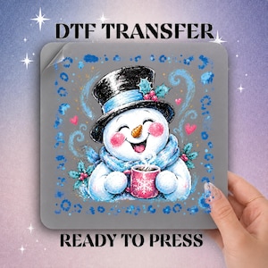 May include: A square DTF transfer sheet featuring a cartoon snowman wearing a black top hat with holly and a blue scarf. The snowman holds a pink mug with a white snowflake design. The background is gray with blue leopard print accents and hearts.