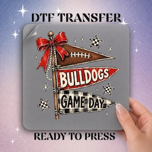 May include: A DTF transfer sheet featuring a graphic of three stacked pennants on a pole with a red bow. The top pennant is brown with a football, the middle is red with the text BULLDOGS, and the bottom is black and white plaid with GAME DAY.