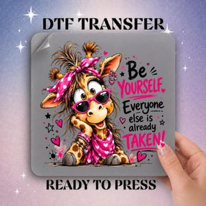 May include: A square DTF transfer featuring a cartoon giraffe wearing pink heart-patterned sunglasses and a matching headscarf. The text reads Be YOURSELF Everyone else is already TAKEN in black and pink lettering, surrounded by small pink hearts.
