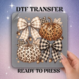 May include: A DTF TRANSFER READY TO PRESS graphic featuring two pumpkins with leopard print patterns, two bows with beige and black buffalo plaid designs, scattered autumn leaves, small acorns, and stars on a gray square background.