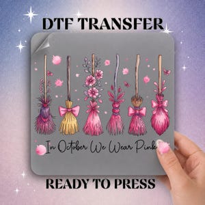 May include: A gray DTF transfer sheet featuring six stylized broomsticks decorated with pink flowers, bows, and butterflies. The text "In October We Wear Pink" is printed in black script below the brooms. The sheet is labeled "DTF TRANSFER" and "READY TO PRESS".