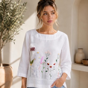 May include: A white long-sleeved linen blouse featuring a delicate, colorful floral embroidery pattern across the front. The design includes various wildflowers in shades of red, pink, yellow, and blue, with green stems and leaves on a plain white fabric.
