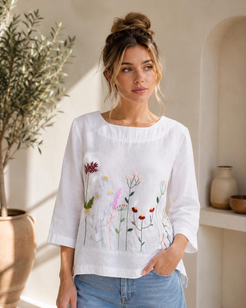 May include: A white long-sleeved linen blouse featuring a delicate, colorful floral embroidery pattern across the front. The design includes various wildflowers in shades of red, pink, yellow, and blue, with green stems and leaves on a plain white fabric.