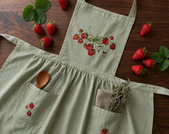 May include: A sage green apron featuring embroidered strawberry vines on the chest and pockets. The apron includes two front pockets, one containing a wooden spoon and metal whisk, the other holding a small bundle of dried herbs.