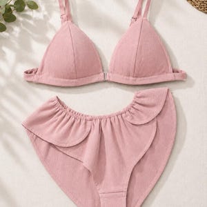 Könnte beinhalten: Ein altrosa Bikini-Set bestehend aus einem Triangel-Oberteil mit dünnen, verstellbaren Trägern und einem Frontverschluss. Das dazu passende Bikini-Unterteil mit hoher Taille ist am oberen Rand mit einem dekorativen Rüschenbesatz versehen.