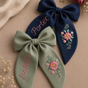 Peut inclure: Deux nœuds pour cheveux en tissu bleu marine et vert sauge. Le nœud bleu marine porte le nom brodé Porter et un motif floral. Le nœud vert sauge porte le nom brodé Isabella et un motif floral assorti avec des fleurs roses et blanches.