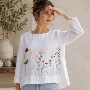May include: A white long-sleeved top featuring a square neckline and colorful floral embroidery across the front. The embroidery depicts various wildflowers in shades of red, pink, yellow, blue, and green. The top is paired with light blue denim jeans.