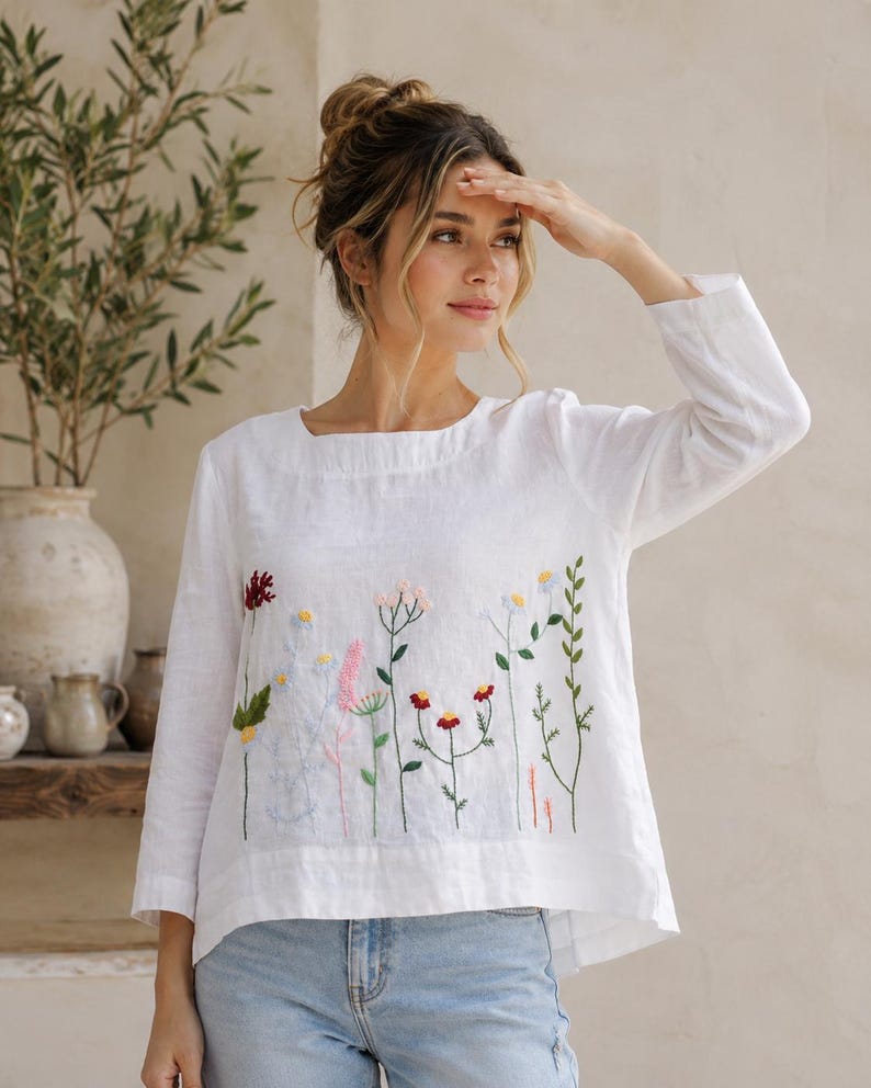 May include: A white long-sleeved top featuring a square neckline and colorful floral embroidery across the front. The embroidery depicts various wildflowers in shades of red, pink, yellow, blue, and green. The top is paired with light blue denim jeans.