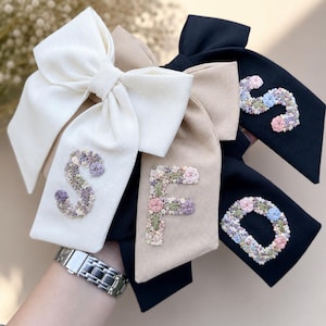May include: Three hair bows in white, beige, and black, each featuring a letter embroidered with small floral patterns. The white bow displays the letter S, the beige bow features the letter F, and the black bow shows the letter D.