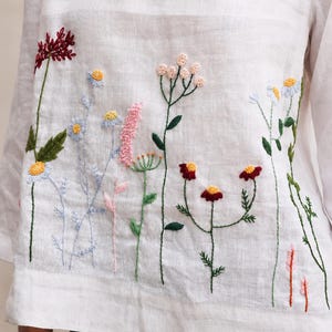 May include: A white long-sleeved shirt featuring colorful floral embroidery across the front. The design includes various wildflowers in shades of red, pink, yellow, and blue with green stems and leaves stitched onto the light-colored fabric.