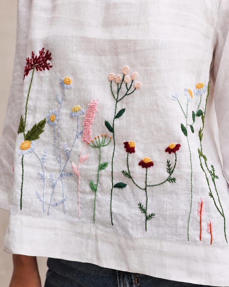 May include: A white long-sleeved shirt featuring colorful floral embroidery across the front. The design includes various wildflowers in shades of red, pink, yellow, and blue with green stems and leaves stitched onto the light-colored fabric.