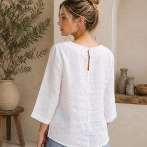 May include: A white linen blouse featuring three-quarter length sleeves, a rounded neckline, and a small button closure at the back. The garment has a straight hemline and a textured fabric finish, paired with blue denim jeans.