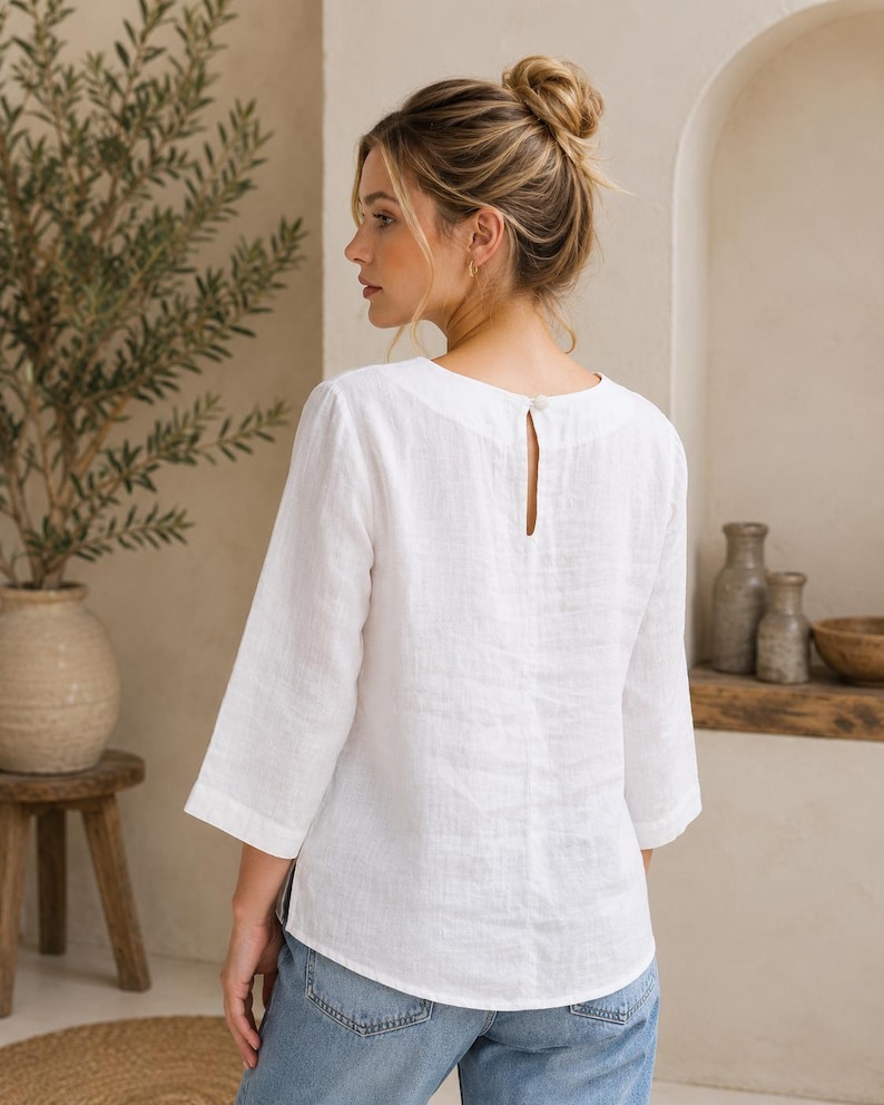 May include: A white linen blouse featuring three-quarter length sleeves, a rounded neckline, and a small button closure at the back. The garment has a straight hemline and a textured fabric finish, paired with blue denim jeans.
