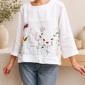 May include: A white long-sleeved top featuring a round neckline and a colorful floral embroidery pattern across the front. The embroidery depicts various wildflowers in shades of red, yellow, pink, and blue with green stems and leaves.