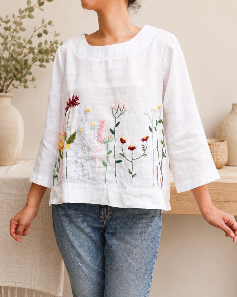 May include: A white long-sleeved top featuring a round neckline and a colorful floral embroidery pattern across the front. The embroidery depicts various wildflowers in shades of red, yellow, pink, and blue with green stems and leaves.