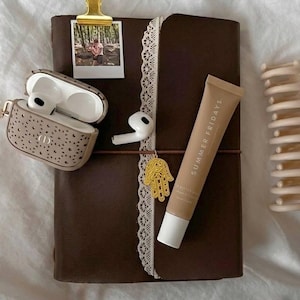 Op de afbeelding: Een donkerbruin lederen dagboek met een witte kanten rand, een gouden metalen clip met een kleine foto, een beige hoesje met patroon voor oordopjes met witte oordopjes, een gouden hamsa-bedel, een tube Summer Fridays lippenbalsem en een beige haarklem.