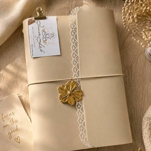 Può includere: Un diario beige con bordo in pizzo bianco, fissato da un elastico con un ciondolo a forma di fiore dorato. Un piccolo cartoncino rettangolare con il testo Inner Lab è fissato in alto. Un foglietto separato riporta la scritta focus on the good.