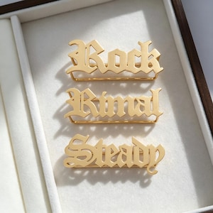 Puede incluir: Tres broches de metal dorado con letras estilizadas en una fuente gótica. El broche superior muestra la palabra Rock, el del medio la palabra Ritual y el inferior la palabra Steady, cada uno con una fina barra horizontal.