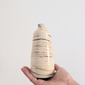 Peut inclure: Un petit vase en céramique avec un col effilé et une base arrondie. La surface présente un émail de couleur crème avec des stries horizontales tourbillonnantes dans des tons gris anthracite foncé et beige clair, créant un aspect marbré.
