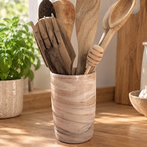 Peut inclure: Un porte-ustensiles cylindrique en céramique avec un motif marbré dans des tons beige, crème et gris doux. Le récipient contient plusieurs ustensiles de cuisine en bois, dont une spatule ajourée, une cuillère pleine et une cuillère à miel.