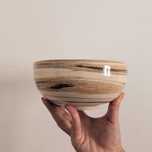 Peut inclure: Un bol en céramique présentant un motif marbré avec des bandes horizontales de couleurs crème, beige et brun foncé. Le bol a une finition lisse et brillante, une forme arrondie avec une base légèrement effilée et un aspect tourbillonnant.
