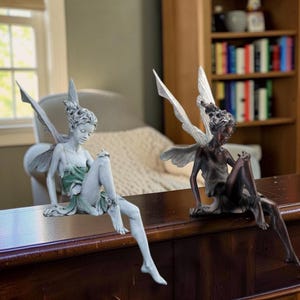 Può includere: Due statuette di fate sedute su un bordo. Una è grigio chiaro con un vestito verde, l'altra è bronzo scuro. Entrambe hanno ali dettagliate, capelli raccolti e sono posizionate con un ginocchio sollevato e una gamba che pende oltre il bordo.