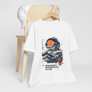 Könnte beinhalten: Ein weißes T-Shirt mit einem Grafikdruck einer Berglandschaft unter einem großen orangefarbenen Mond, mit Kiefern, Wellen und dem Text "MIDNIGHT MOUNTAIN CLUB" in schwarzer Schrift. Eine weiße Baseballkappe liegt auf der hölzernen Stuhllehne.
