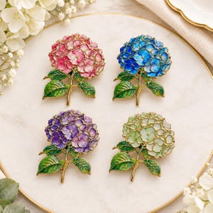May include: Four hydrangea flower brooches with gold-tone metal outlines. Each brooch features enamel petals in distinct colours: pink, blue, purple, and pale green, all attached to a gold-tone stem with two detailed green leaves at the base.