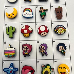 May include: A collection of twenty colourful silicone shoe charms arranged in a grid, featuring various pop culture characters, food items, emojis, and symbols including a Pokeball, a star, a cactus, a mushroom, and a SpongeBob character.