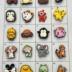 May include: A collection of twenty colourful rubber shoe charms arranged in a grid, featuring various cartoon characters, animals, and objects including a bear, penguin, Pikachu, dog, mushroom, koala, Charmander, bee, and a duck holding a knife.