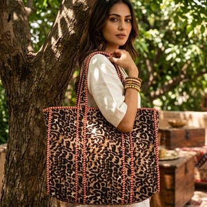 Puede incluir: Un bolso tote rectangular grande con un estampado de leopardo en tonos marrones y negros sobre tela texturizada. El bolso incluye dos correas largas para el hombro y un ribete trenzado rojo y blanco en los bordes y costuras.