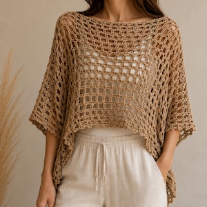Op de afbeelding: Een beige gehaakte poncho met een open vierkant rasterpatroon, een brede boothals en korte mouwen. Het kledingstuk wordt gedragen over een effen wit hemd en een lichtgekleurde broek met een trekkoord in de taille.