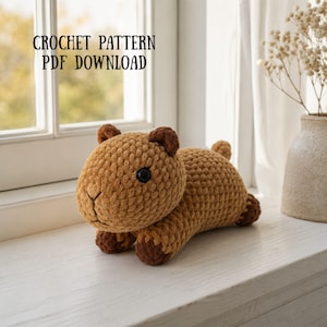 Puede incluir: Un juguete de peluche de capibara tejido a ganchillo con hilo marrón claro y detalles en marrón oscuro en las orejas y patas, con ojos de plástico negro y nariz bordada. El texto CROCHET PATTERN PDF DOWNLOAD aparece en la esquina superior izquierda.