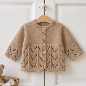 Könnte beinhalten: Eine beigefarbene, langärmelige Strickjacke mit Rundhalsausschnitt, fünf hellen Knöpfen in der Mitte und einem dekorativen Zickzack-Lochmuster am unteren Teil des Kleidungsstücks sowie an den Ärmelbündchen.