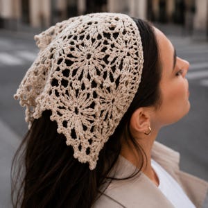 May include: A beige crochet headscarf featuring a repeating floral motif with intricate openwork patterns. The triangular-shaped accessory is crafted from fine yarn, showcasing a delicate, lacy texture that covers the top of the head.