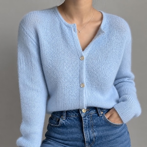 Può includere: Un cardigan a maniche lunghe color azzurro con scollo rotondo e chiusura frontale con tre bottoni madreperlacei visibili. Il capo in maglia presenta una consistenza morbida e soffice ed è abbinato a un paio di jeans blu in denim.