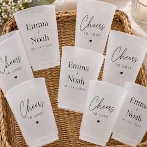 May include: A set of seven frosted plastic cups featuring black printed text. Some cups display the names Emma and Noah with the date 14 | 06 | 2027, while others feature the phrase Cheers to Love with a small heart icon below the text.