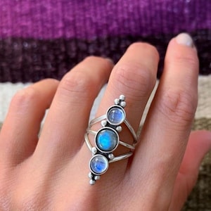 Puede incluir: Un anillo de plata con tres piedras de luna redondas e iridiscentes dispuestas verticalmente. Las piedras muestran un brillo azul, engastadas en una banda de metal pulido que se divide en varios hilos, acentuada por pequeñas esferas de plata.