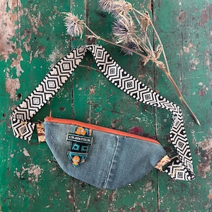 Könnte beinhalten: Eine Bauchtasche aus Denim mit orangefarbenem Reißverschluss und einem schwarz-weiß gemusterten Riemen. Die Tasche hat einen kleinen dekorativen Aufnäher mit orangefarbenen Blumenakzenten und ein rechteckiges Metallschild mit dem Text CRAVATEUR.