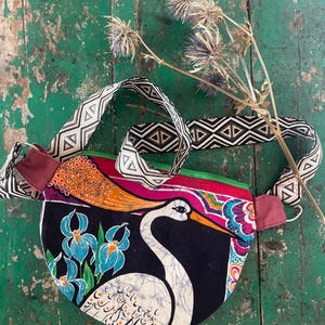 Könnte beinhalten: Eine halbmondförmige Umhängetasche mit einer weißen Kranich-Illustration auf schwarzem Grund und blauen Irisblüten. Die Tasche hat einen leuchtend bunten oberen Rand und einen breiten, verstellbaren Riemen mit schwarz-weißem Rautenmuster.