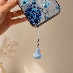 Könnte beinhalten: Eine transparente Smartphone-Hülle mit einer blauen Meeresschildkröte und Muschelgrafik, befestigt an einer hellblauen Kordel mit einem kleinen, hellblauen, perlenbesetzten Entenanhänger, einem leuchtend orangefarbenen Schnabel und einem silbernen Karabiner.