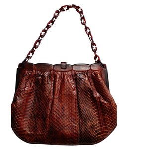 May include: A dark reddish-brown handbag featuring a textured snakeskin pattern. The bag has a structured top frame and a matching dark reddish-brown chain-link handle. The body of the bag is gathered, creating a pleated effect below the frame.