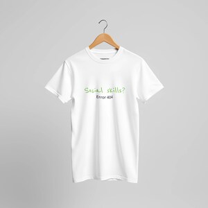 Puede incluir: Una camiseta blanca de manga corta con el texto Social skills? en fuente manuscrita verde y el texto Error 404 en fuente sans-serif negra centrado en el pecho. La prenda se muestra en una percha de madera sobre un fondo claro y liso.
