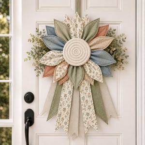 Puede incluir: Un adorno de tela para puerta con un disco central acolchado rodeado de p&eacute;talos superpuestos en tonos azul apagado, verde salvia, terracota y estampados florales. El dise&ntilde;o incluye follaje y largas cintas de tela puntiagudas que cuelgan.