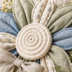 Puede incluir: Un adorno de pared con forma de flor de tela con p&eacute;talos acolchados en tonos verde salvia, azul polvoriento y crema con estampados florales. El centro es un disco de tela enrollado de color crema con costuras visibles y un lazo de lino.