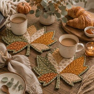 Puede incluir: Dos posavasos de tela acolchada con forma de hojas de arce, con un dise&ntilde;o de patchwork en tonos verdes, naranjas y crema con patrones florales y de cuadros. Cada posavasos sostiene una taza de cer&aacute;mica con una bebida marr&oacute;n claro.