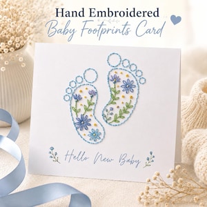 May include: A white greeting card featuring hand-embroidered baby footprints outlined in blue thread. The footprints are filled with floral embroidery in shades of blue, green, and yellow. The text "Hello New Baby" is printed in blue script below.