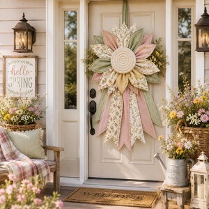 Puede incluir: Una corona de flores de tela colgada en una puerta clara, con p&eacute;talos superpuestos en tonos verde salvia, rosa empolvado y estampados florales amarillos, un disco central de cuerda trenzada y largas cintas de tela puntiagudas.