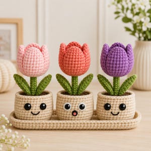 May include: Three crocheted tulips in light pink, coral, and purple, each planted in a beige crocheted pot with a unique embroidered face. The pots sit on a matching beige crocheted rectangular tray, creating a cohesive set of soft textile decor.